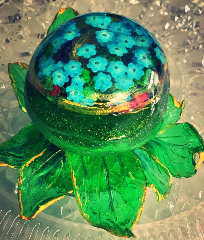 A translucent green sugar paperweight featuring a spherical top with embedded blue sugar flowers, resting on a base of sculpted green sugar leaves with gold accents.