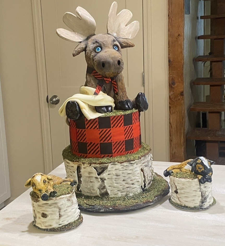 A woodland-themed cake set featuring a two-tiered cake with a moose figurine topper on a red and black buffalo plaid upper tier, birch bark-textured lower tier, accompanied by two smaller cakes each adorned with a lamb and goat figurine in grassy settings.