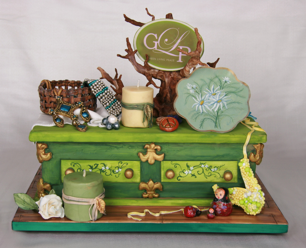An elaborate green fondant cake designed to look like a vintage chest of drawers, decorated with gold accents, a tree branch topper with a logo, and various accessories including a basket, candles, and a painted plate.