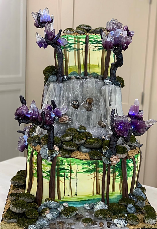 This elaborate two-tiered cake is decorated with a painted forest scene, faux moss, rocks, and purple crystal clusters, featuring a central waterfall cascading down the middle tier.