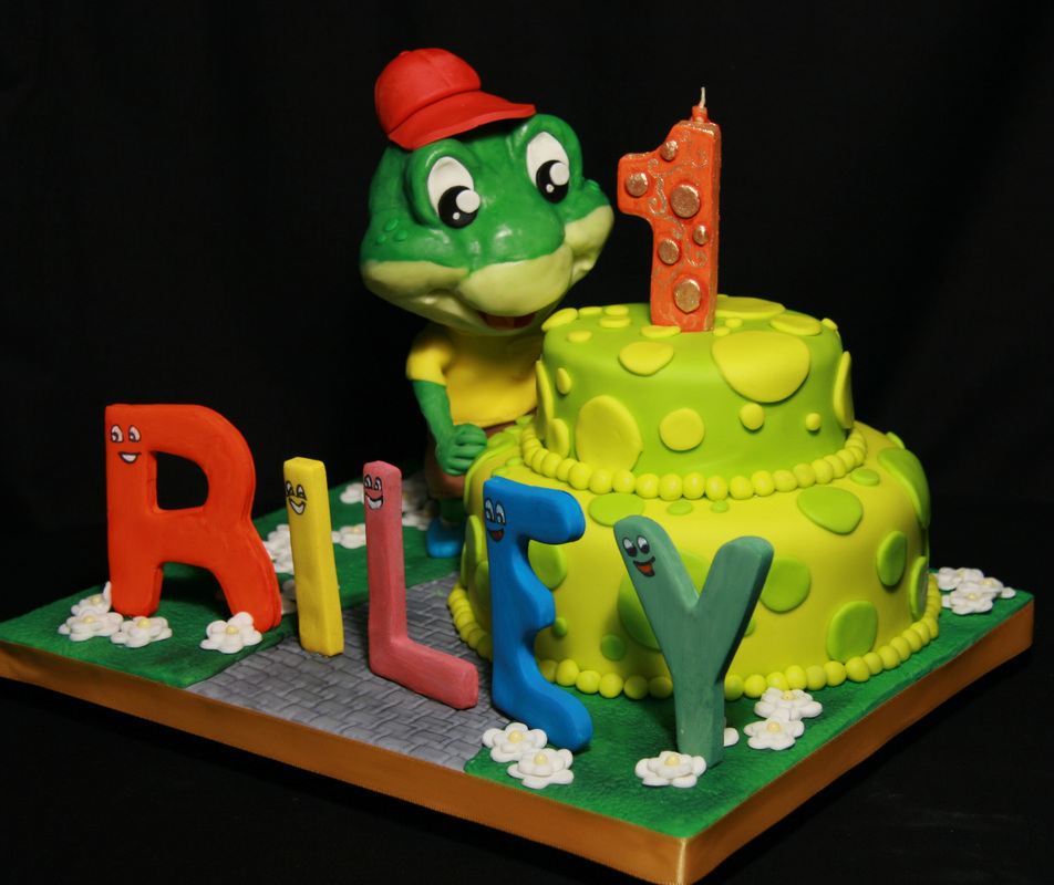 A two-tiered lime green birthday cake decorated with a large orange number one candle and a frog mascot wearing a red cap, accompanied by colorful fondant letters spelling "RILEY" on a grassy base.