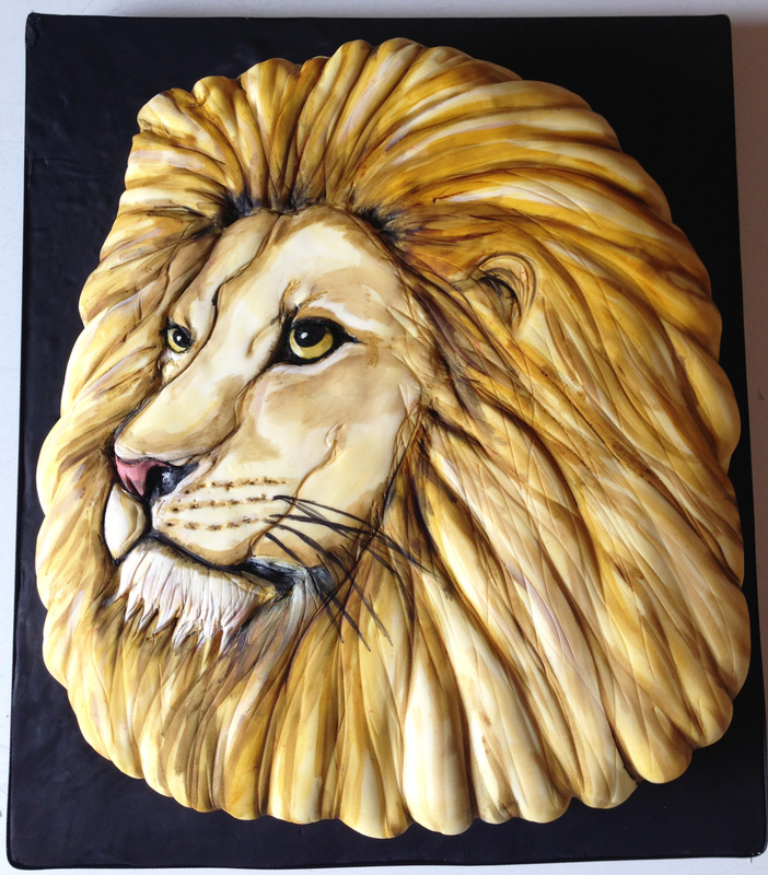 A realistic lion head cake with a sculpted, textured mane in shades of golden brown and cream, featuring detailed painted facial features including amber eyes, a pink nose, and black whiskers, set against a black background.