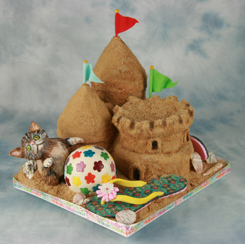 A detailed cake designed to look like a sandy beach scene features a sandcastle with three flags, a realistic cat figurine, a colorful beach ball, a towel, and flip-flops.