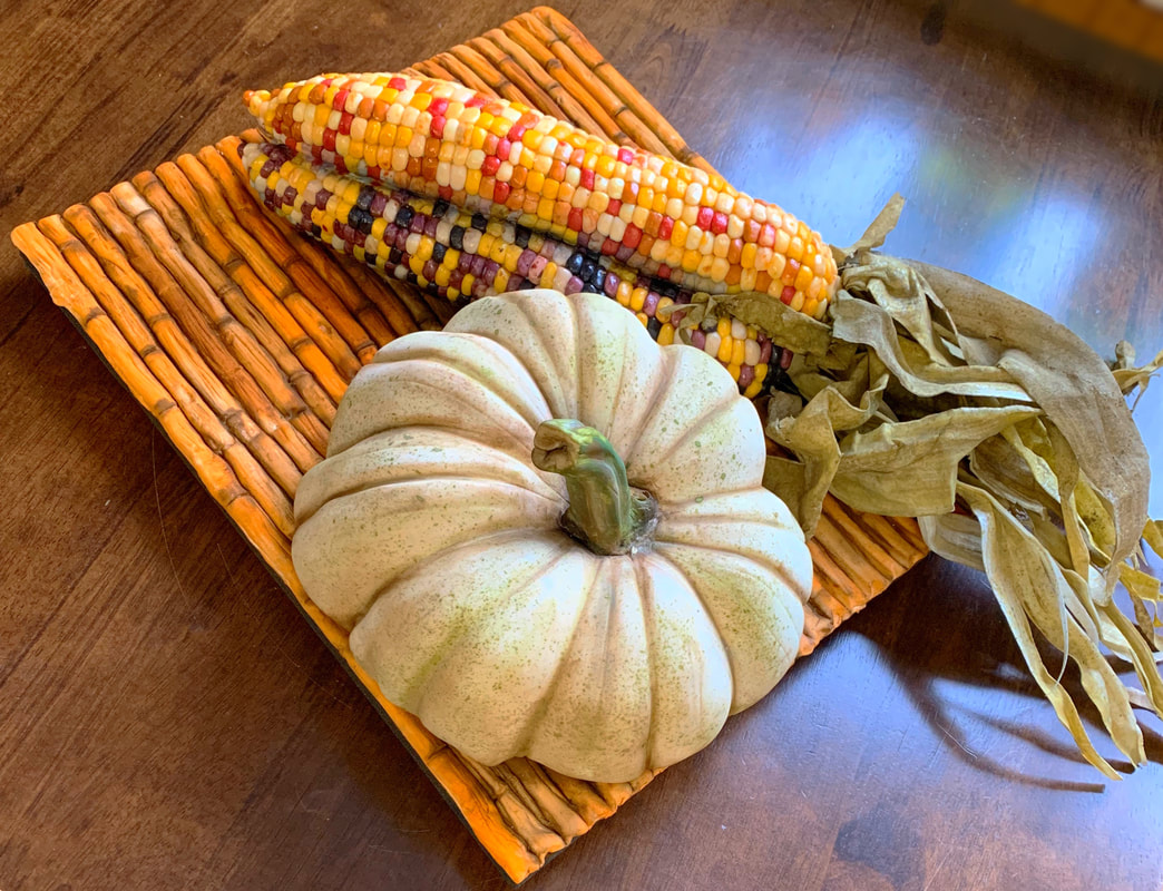 A small, pale green and white pumpkin rests on a woven orange placemat alongside two ears of colorful Indian corn with yellow, red, and purple kernels.