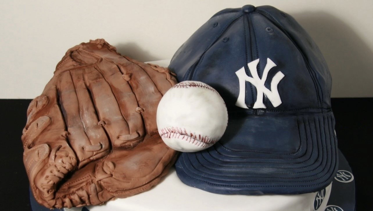 A sculpted cake featuring iconic New York Yankees baseball memorabilia — a brown leather-style baseball glove, a white baseball with red stitching, and a navy blue Yankees cap with the classic interlocking "NY" logo — all resting atop a smooth white-frosted cylindrical cake base with repeating Yankees logos.