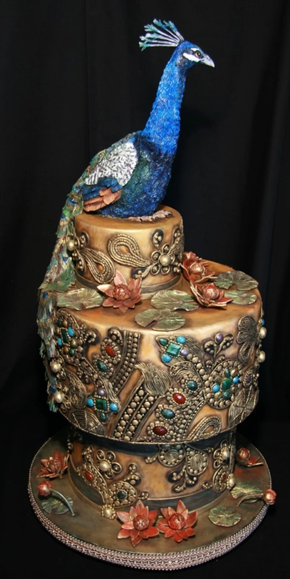 An ornate, multi-tiered cake with gold and bronze filigree detailing and jeweled accents is topped with a vibrant blue peacock figurine.
