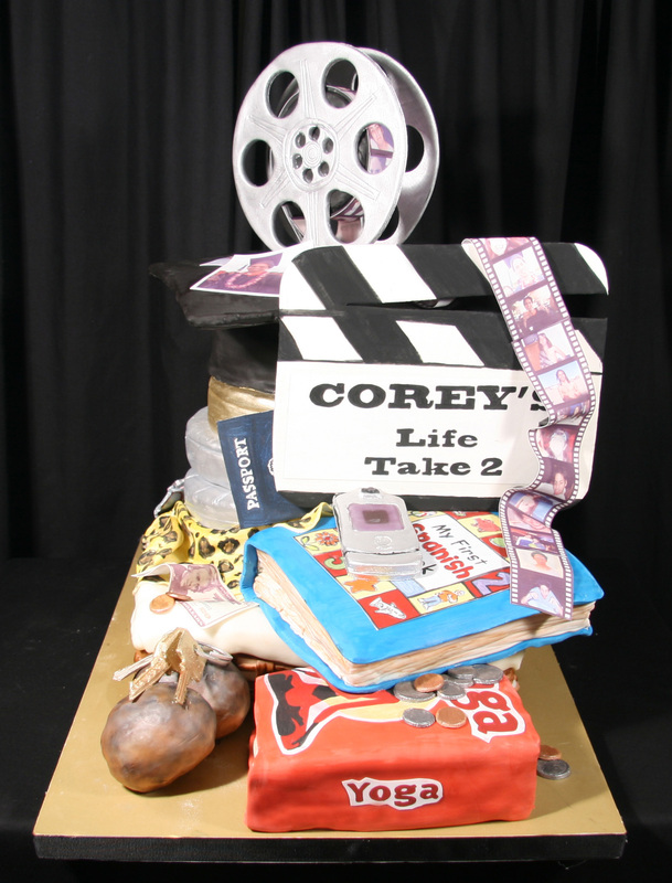 A multi-tiered cake decorated with film-themed elements, including a silver film reel, a clapperboard reading "COREY'S Life Take 2," a blue book, a passport, a mobile phone, and a red block labeled "Yoga."