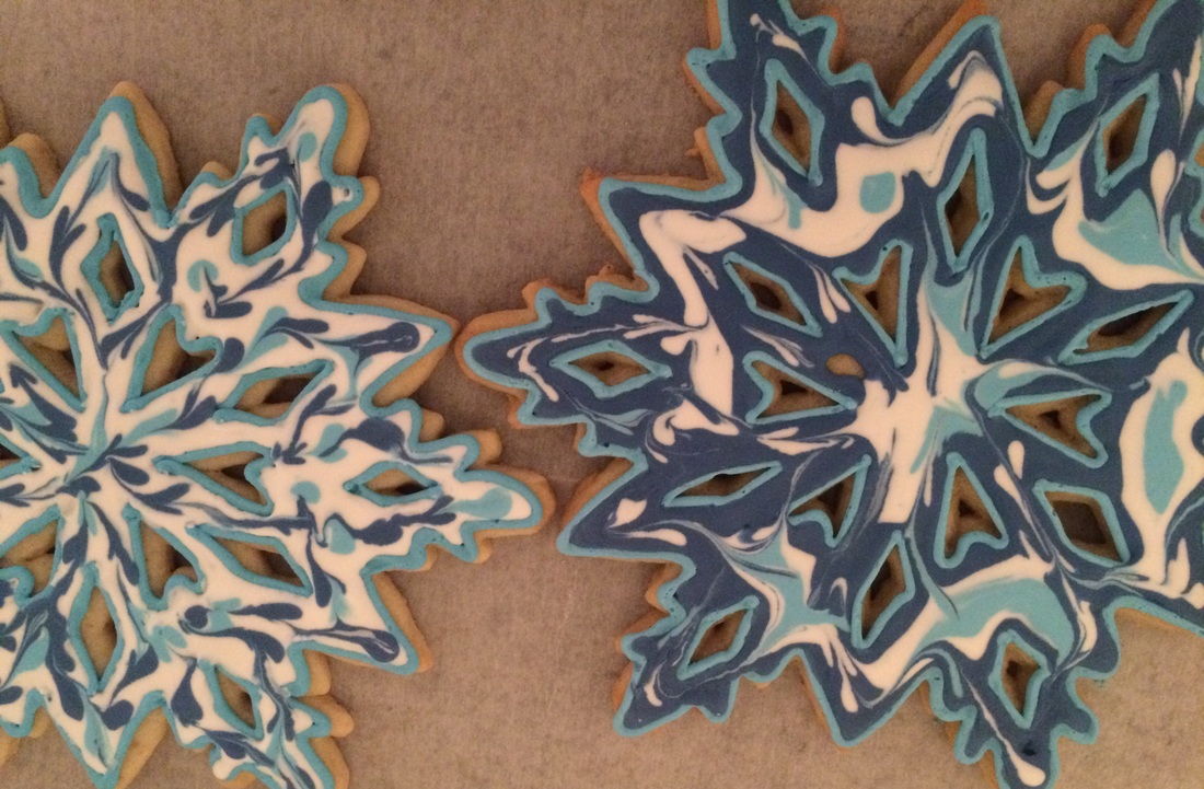 Two intricately decorated snowflake-shaped sugar cookies feature a marbled icing design in white, light blue, and dark blue, resting on a brown parchment paper surface.