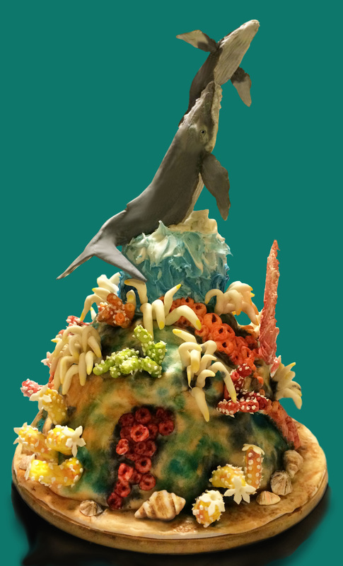 A sculpted cake designed to resemble an underwater coral reef, featuring two grey whales leaping from a blue frosting wave at the top, surrounded by colorful coral made from cereal and candy.