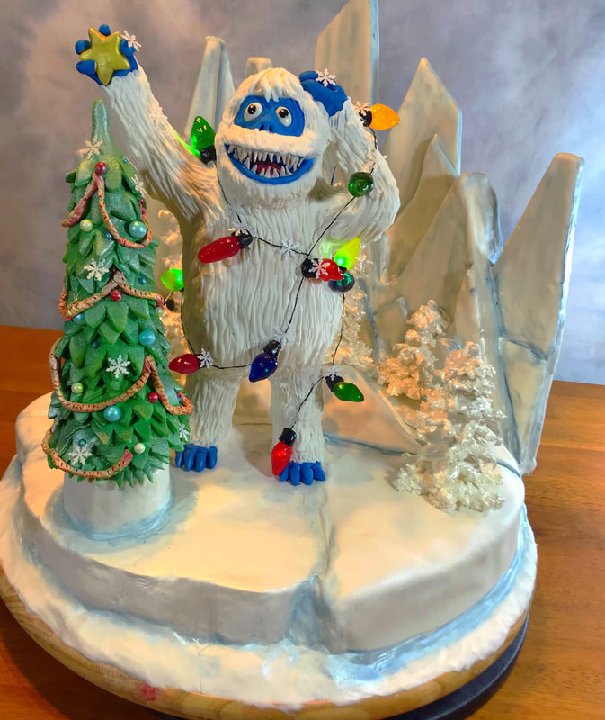 A whimsical, sculpted cake features a large, white-furred, blue-faced monster character joyfully holding a star-topped Christmas tree and wrapped in colorful Christmas lights, set against a backdrop of icy blue mountains and a snowy landscape.