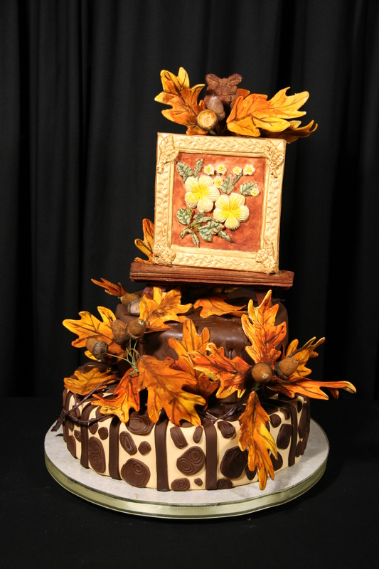 A two-tiered chocolate cake decorated with autumn-themed fondant leaves, acorns, and a framed floral painting topper sits on a marble cake board against a black backdrop.