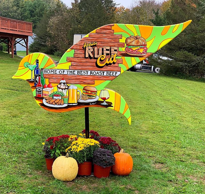 A colorful, bird-shaped wooden sign for "Annie's Rustic Cut," advertising "Home of the Best Roast Beef" with illustrations of food and drinks, stands on a grassy lawn surrounded by potted mums and pumpkins.