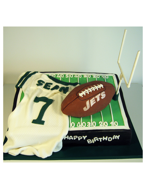 A rectangular birthday cake decorated with a green football field design, topped with a white jersey reading "SEAN 7," a brown football labeled "JETS," and a white goalpost, with "HAPPY BIRTHDAY" written in white icing on the black side.