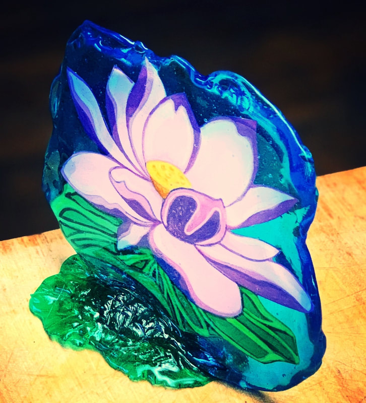 A translucent blue and green sugar sculpture featuring a painted white and purple lotus flower with a yellow center, resting on a wooden surface.