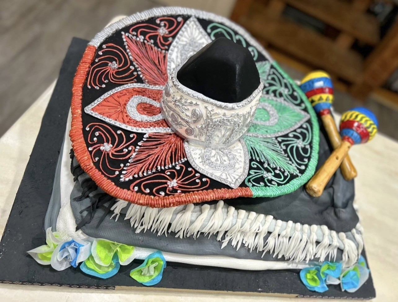 A Mexican-themed cake featuring a vibrant sombrero as centerpiece with wide brim adorned in bold red, green, and silver patterns against black, ornate silver detailing on the crown, white fondant tassels mimicking fringe, two colorful maracas in yellow, blue, and red, and delicate sugar flowers in blue, green, and white.