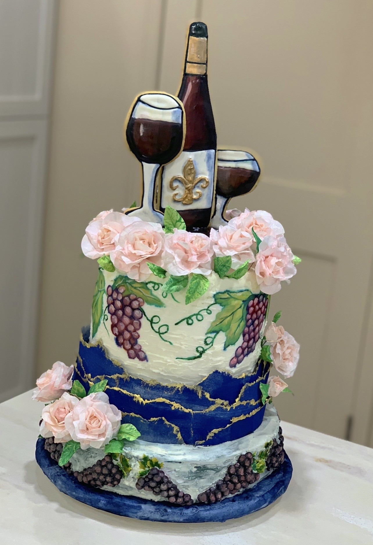 A three-tiered wine-themed cake — top tier in creamy white buttercream hand-painted with purple grapes and green vines, crowned by blush-pink sugar roses and wine bottle with two glasses topper, middle tier in deep cobalt blue with gold leaf kintsugi design, bottom tier in marbled gray-blue with piped dark grape clusters and blush roses.