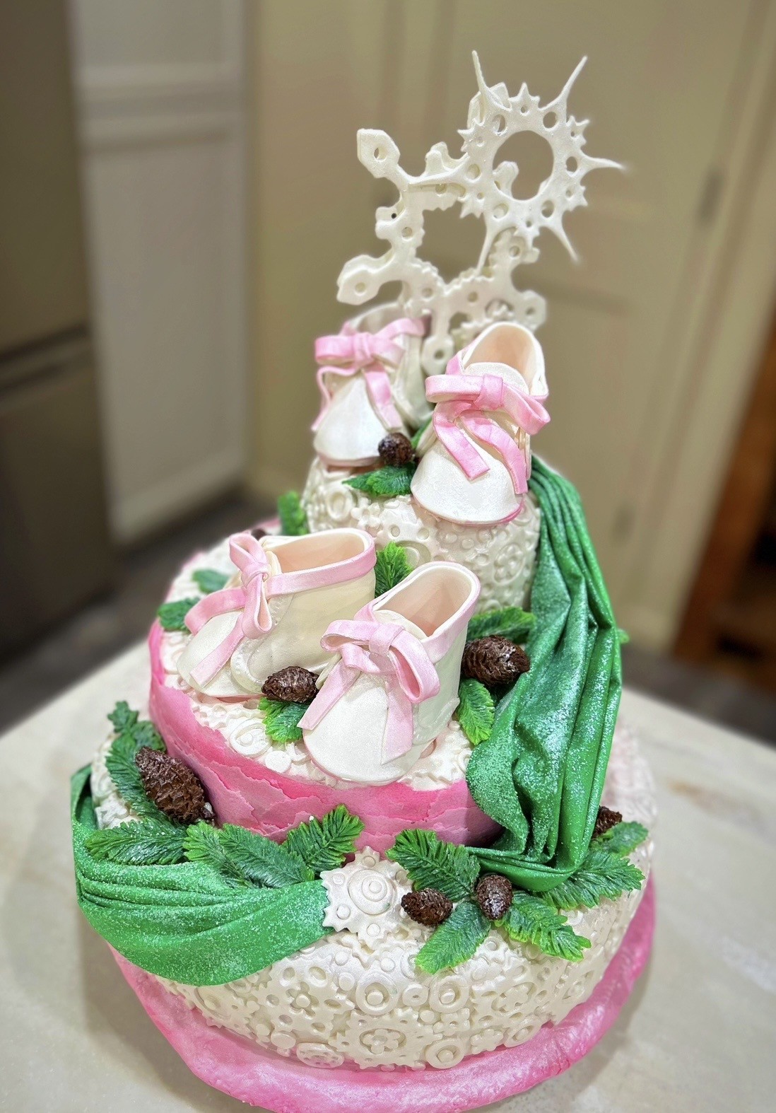 A three-tiered cake with soft pink fondant base featuring white lace-like patterns and a shimmering green ribbon cascade intertwined with lifelike green leaves and pinecone decorations, middle tier adorned with miniature fondant baby shoes with pink bows, top tier with another set of baby shoes and an elaborate white sugarwork crown topper.