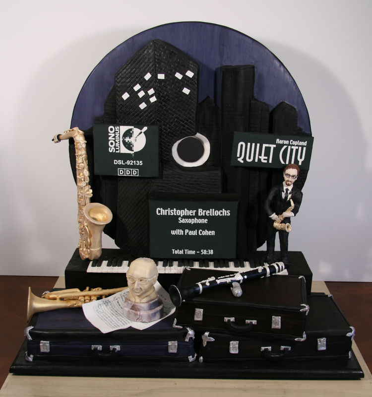 An elaborate black and blue fondant cake decorated with a cityscape backdrop, featuring edible replicas of musical instruments like a saxophone, clarinet, and trumpet, alongside printed album covers and a figurine of a saxophonist.