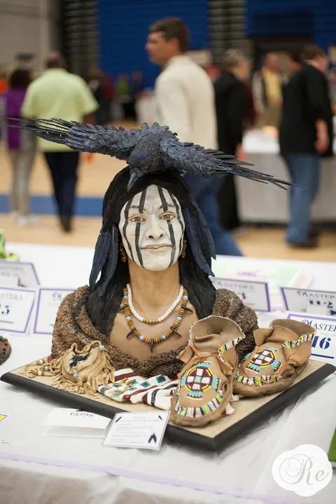 A hyper-realistic cake sculpture depicts a Native American figure with black and white face paint, a raven perched on their head, and beaded moccasins arranged on the display board.