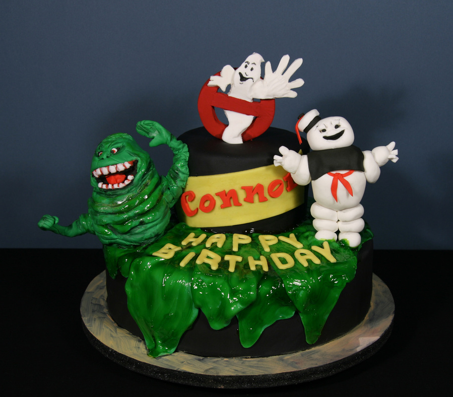 A two-tiered black birthday cake decorated with fondant figures of Slimer and the Stay Puft Marshmallow Man, topped with the Ghostbusters logo, and featuring the name "Connor" and "Happy Birthday" amidst green slime.