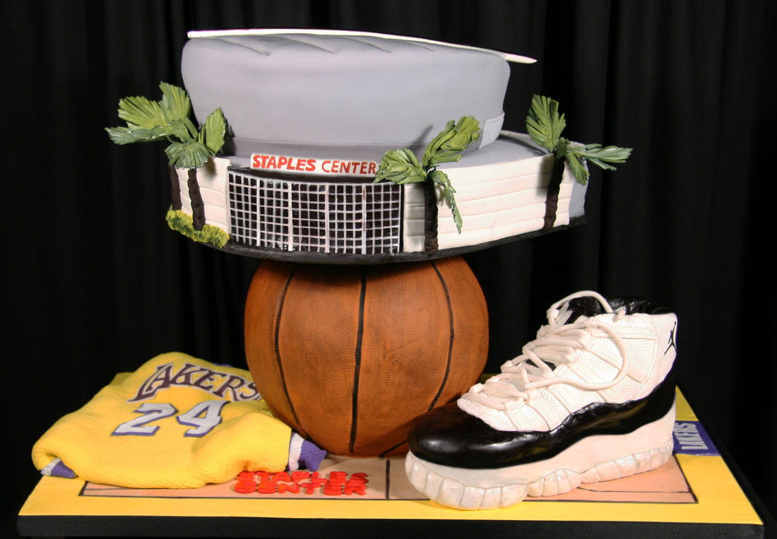 A detailed cake sculpture features a replica of the Staples Center arena perched atop a basketball, accompanied by a Lakers jersey with the number 24 and a sneaker, all set on a court-themed base.