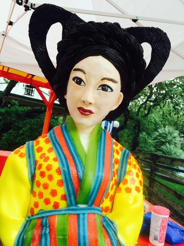 A colorful sugar sculpture of a woman in traditional Japanese attire, featuring a vibrant yellow kimono with red floral patterns, a green collar, and a striped obi, topped with an elaborate black hairstyle, set against a backdrop of greenery and a tent structure.