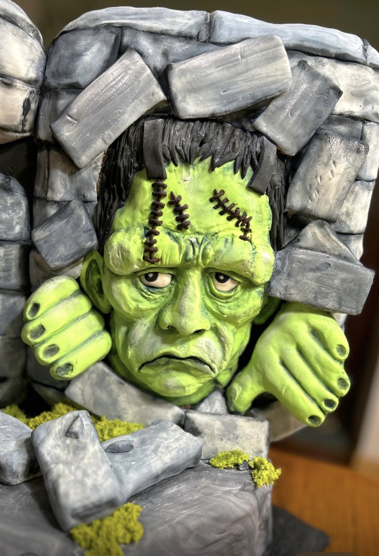A sculpted cake designed as Frankenstein's monster emerging from a rocky tomb with gray stone blocks, the creature's green stitched face and hands bursting through, furrowed brow, sunken eyes, downturned mouth, knotted black hair, articulated fingers gripping the stones, with mossy yellow-green foreground.
