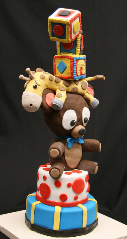 A whimsical, multi-tiered cake features a blue and yellow striped base, a white tier with red polka dots, a large brown teddy bear wearing a blue bow tie, a yellow giraffe resting on the bear's head, and a stack of colorful toy blocks balancing on top.