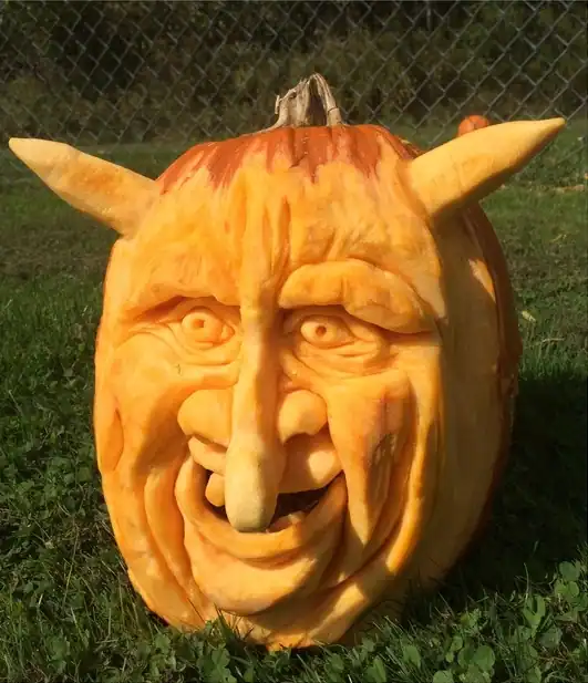 A detailed vegetable carving of a devilish face with horns, a long nose, and a wide grin sits on a grassy lawn in front of a chain-link fence.