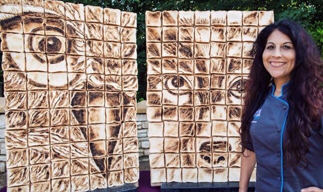 A smiling woman with long dark hair stands beside two large vertical panels of toasted marshmallows arranged to create pixelated mosaic portraits of an owl and a dog.