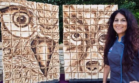 A smiling woman with long dark hair stands beside two large vertical panels of toasted marshmallows arranged to create pixelated mosaic portraits of an owl and a dog.