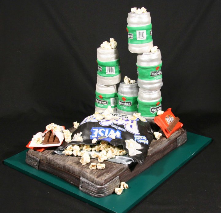A novelty cake designed to look like a wooden crate overflowing with a bag of Wise potato chips and Kit Kat bars, with five fondant Heineken beer cans stacked in a pyramid formation and topped with popcorn.