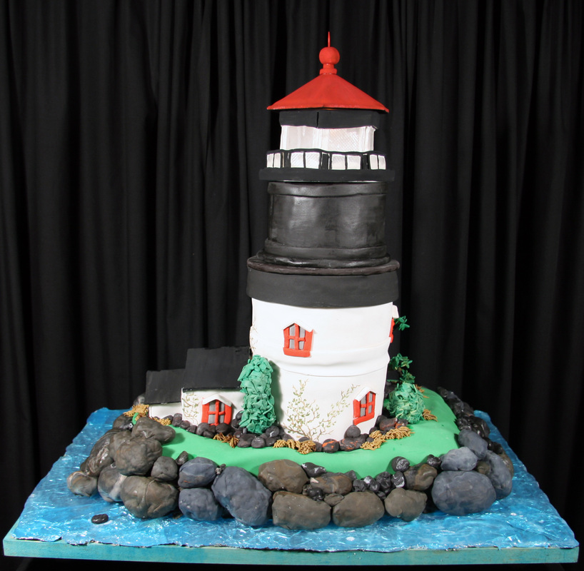 A detailed lighthouse cake featuring a white tower with black bands, a red roof, and small red windows, set on a green grassy base surrounded by gray fondant rocks and blue icing representing water.