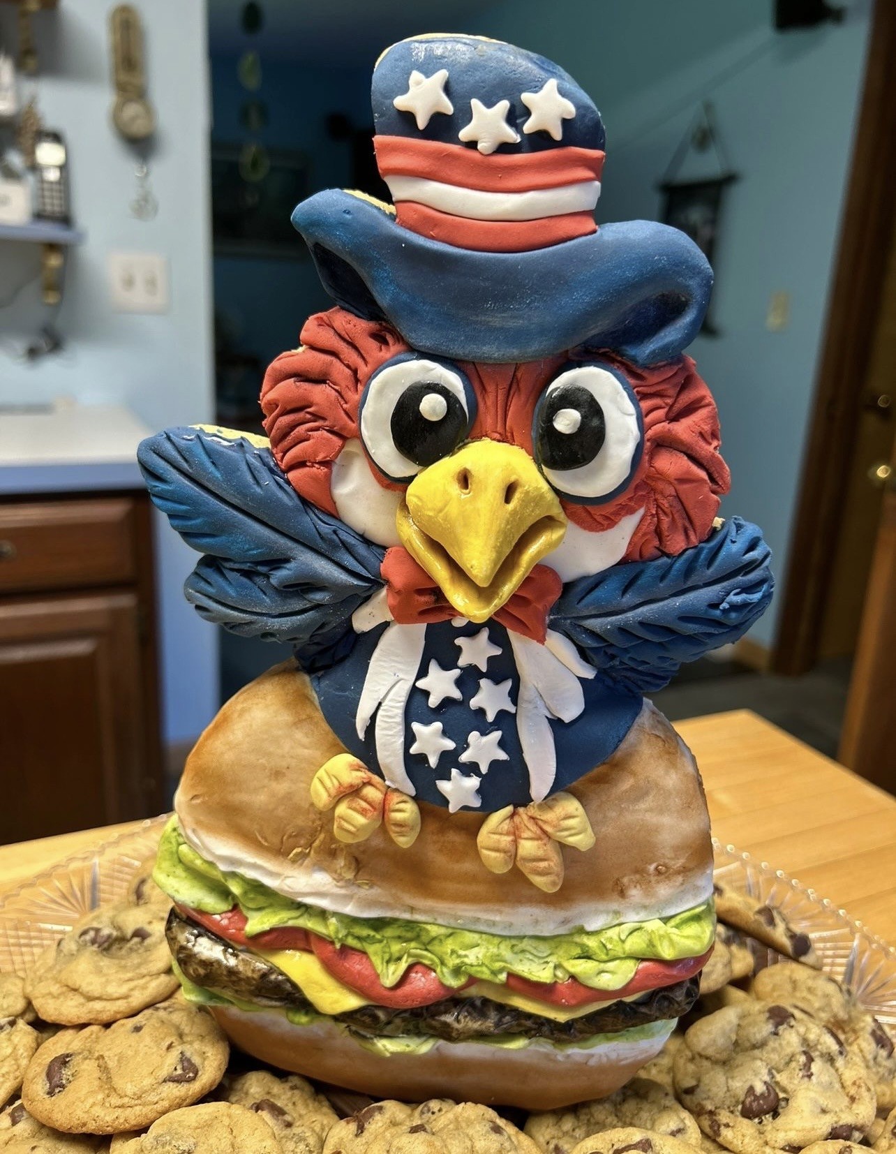 A patriotic sculpted cake featuring a cartoonish bald eagle perched atop a realistic hamburger, the eagle wearing a blue top hat with white stars and red-and-white stripes, blue vest patterned with white stars, deep blue wings with textured feather details, talons gripping the burger bun, surrounded by chocolate chip cookies.