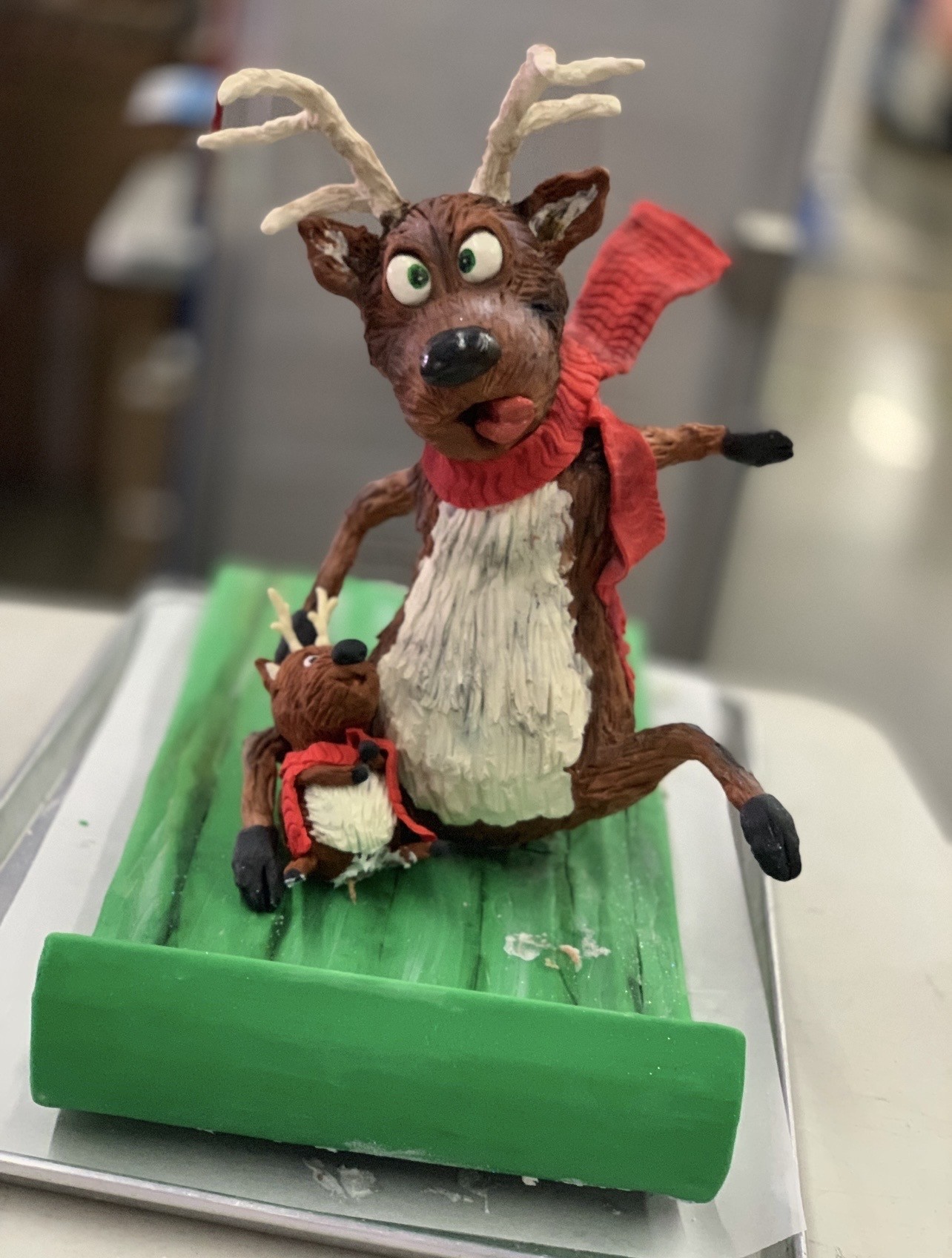 A sculpted cake diorama featuring two anthropomorphic reindeer on a bright green sled — the larger reindeer mid-action with one leg kicked up, wide green eyes, prominent black nose, red scarf flowing behind, and the smaller reindeer looking up in delight, both wearing red scarves with small branching antlers.