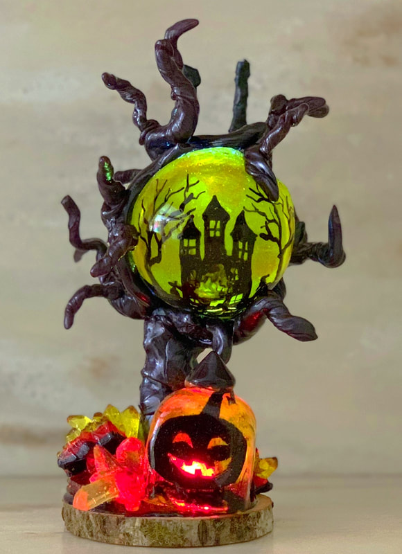 A Halloween-themed sugar figurine features a glowing green crystal ball with a haunted house silhouette, held by twisted dark sugar branches, and sits above an illuminated jack-o'-lantern on a wooden base surrounded by colorful sugar gems.