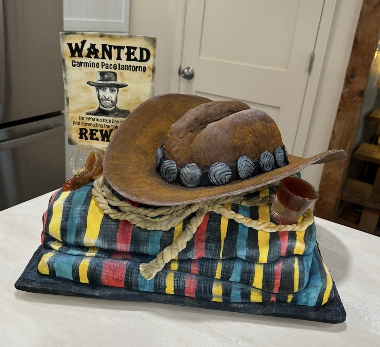 A rustic Western-themed cake featuring a large realistic cowboy hat crafted from fondant resting atop a stack of colorful striped fabric resembling a blanket or saddle pad, with a coiled twine rope, layered blocks in vibrant stripes of yellow, red, blue, and black, a translucent glass of amber liquid, and a "WANTED" poster for "Carmine Pace Iantorno."