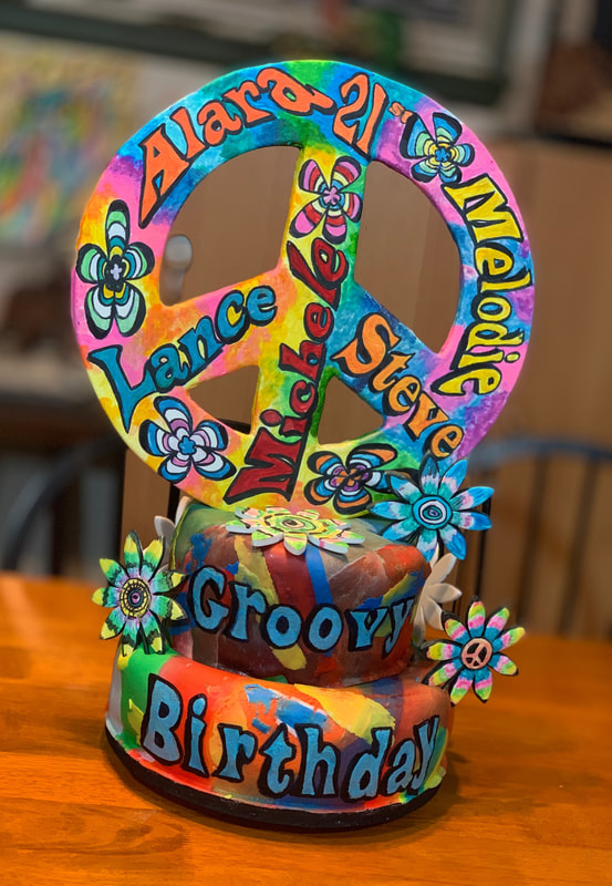 A psychedelic, tie-dye themed birthday cake features a large peace sign topper with names and the phrase "Groovy Birthday" written in colorful lettering.