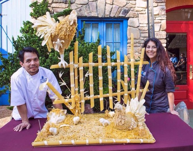Two chefs stand behind an elaborate food sculpture made of yellow ingredients, featuring a rooster perched on a bamboo fence above a hen in a nest.