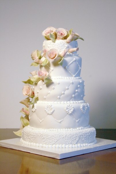 A four-tiered white wedding cake decorated with intricate piped icing and adorned with a cascade of pale pink roses and green leaves.