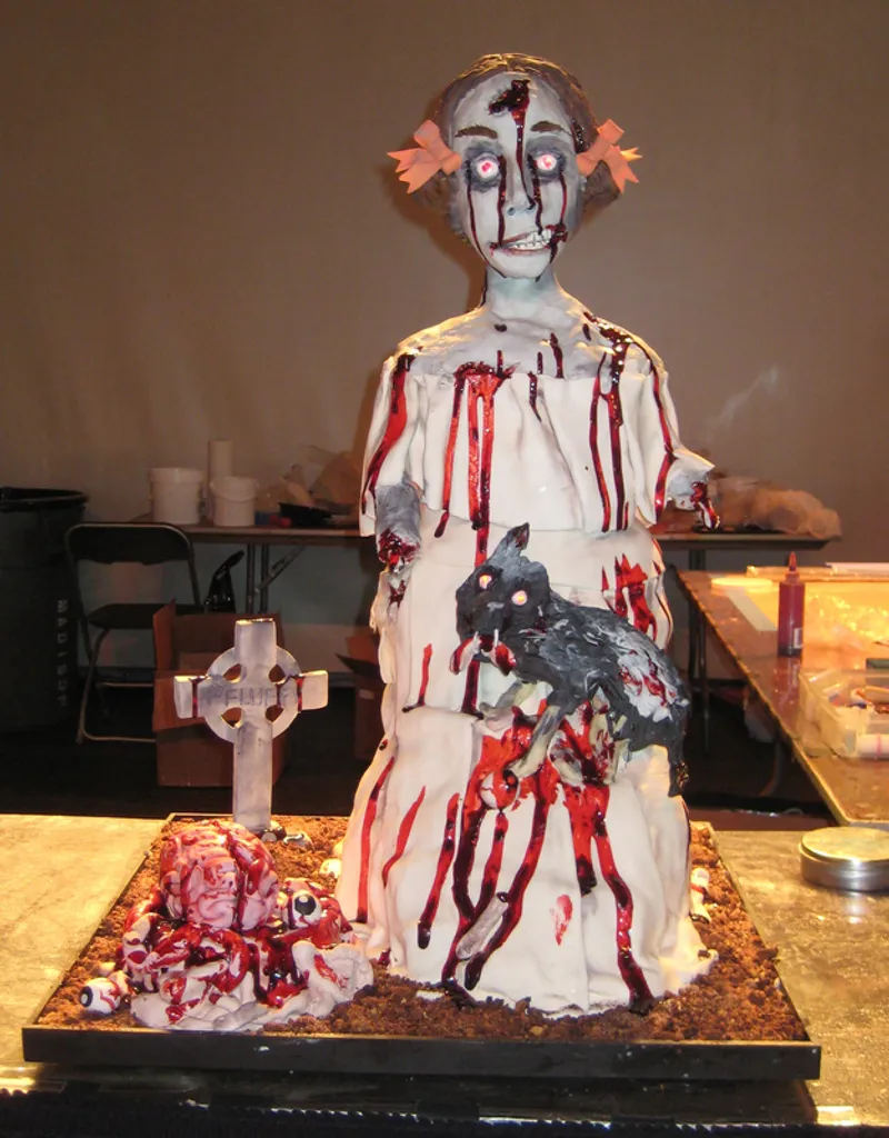 A gruesome, multi-tiered cake sculpted to resemble a bloody zombie girl in a torn white dress, holding a black wolf, with a gory pile of flesh and eyeballs at her feet next to a small Celtic cross.