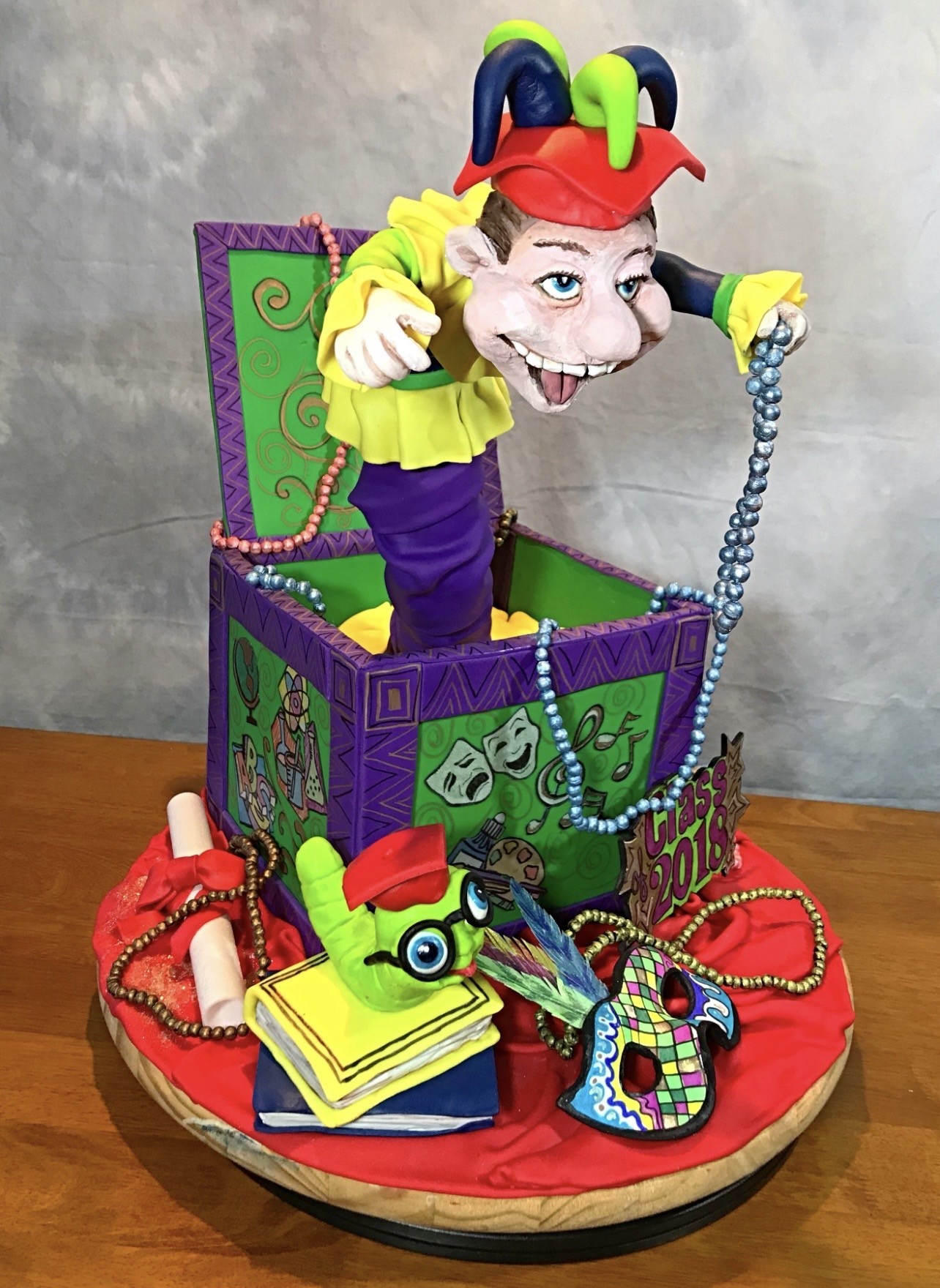 A theatrical jack-in-the-box cake featuring a jester character in a red hat with blue and green horns emerging from a purple and green box adorned with theater masks, musical notes, and abstract patterns, surrounded by fondant props including a feathered mask, books topped with a quirky hand wearing glasses, a scroll, and beads, on a red fondant stage curtain base with a "Class of 2018" plaque.