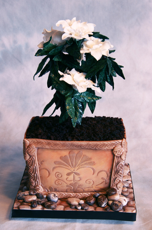 An elaborate cake sculpted to resemble a potted plant features white flowers and green leaves rising from dark crumb "soil" in a decorative tan planter, surrounded by small fondant rocks on a black base.