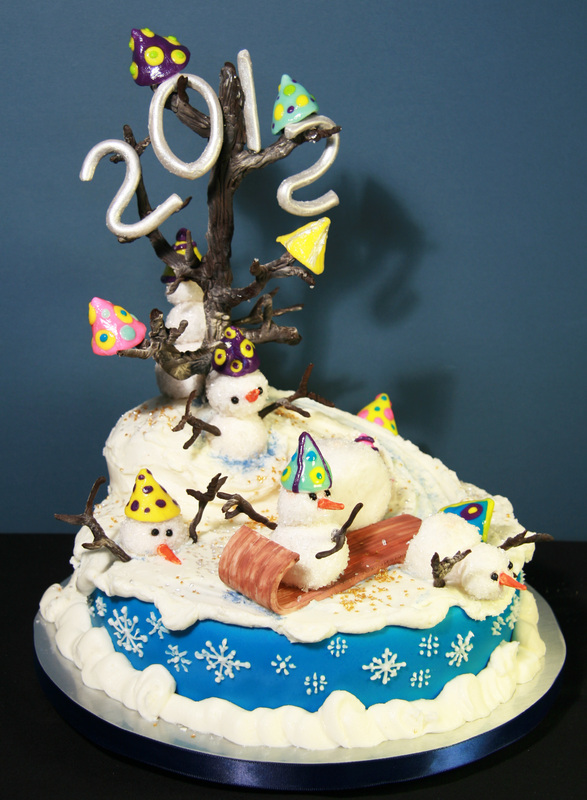 A festive 2012 New Year's cake decorated with a blue base, white snow, and multiple snowmen wearing colorful party hats, topped with a branch tree displaying the year "2012" in white numerals.