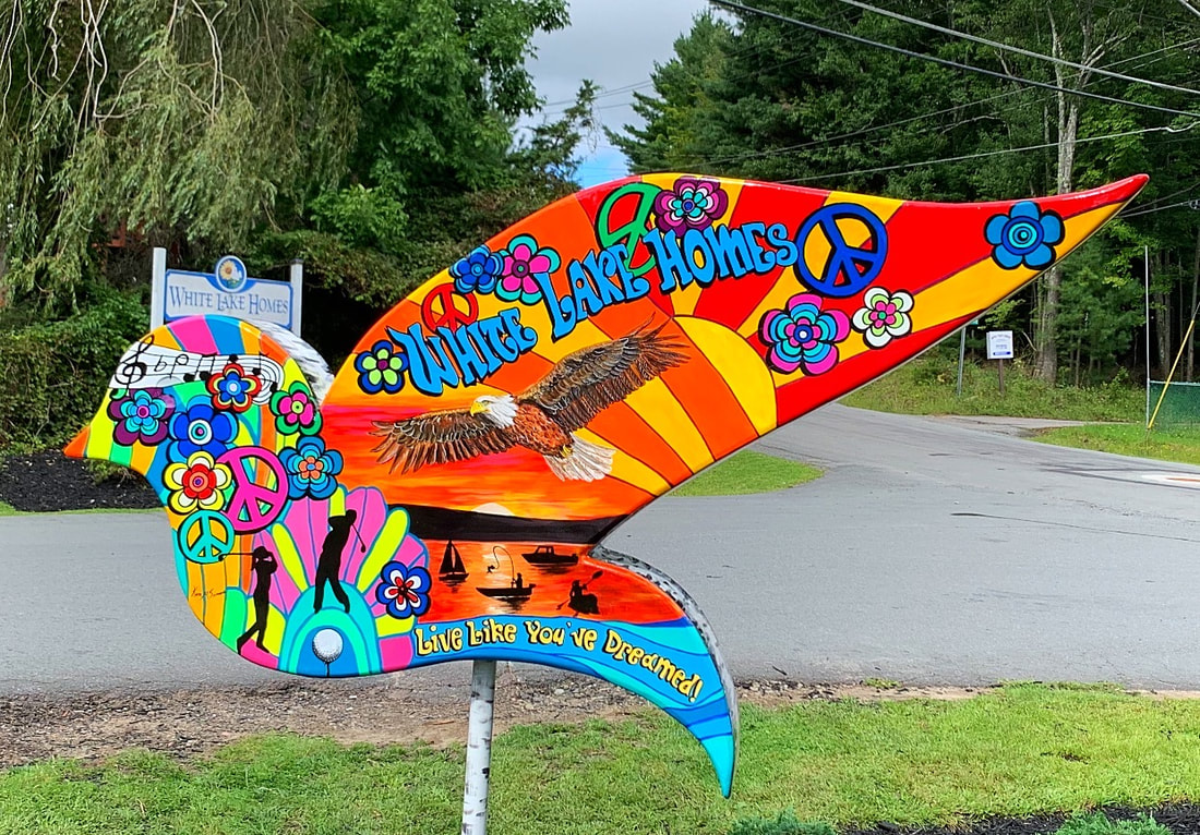 A colorful, psychedelic-style bird-shaped sign for "White Lake Homes" features peace symbols, flowers, a bald eagle, and the slogan "Live Like You've Dreamed!"