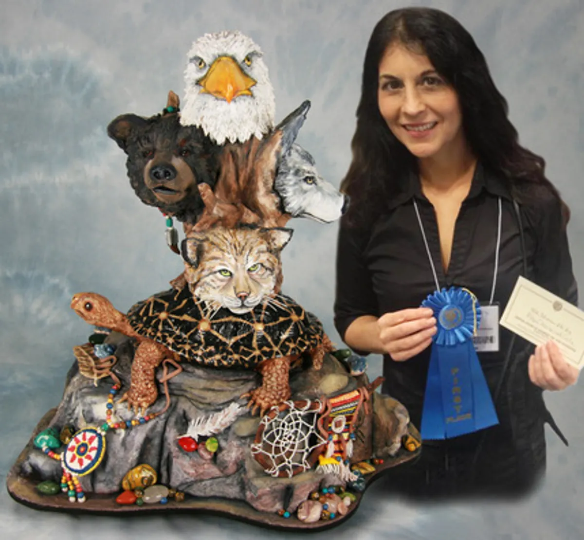 Kim Simons holding a blue ribbon and certificate beside a cake decorated with stacked woodland animals.