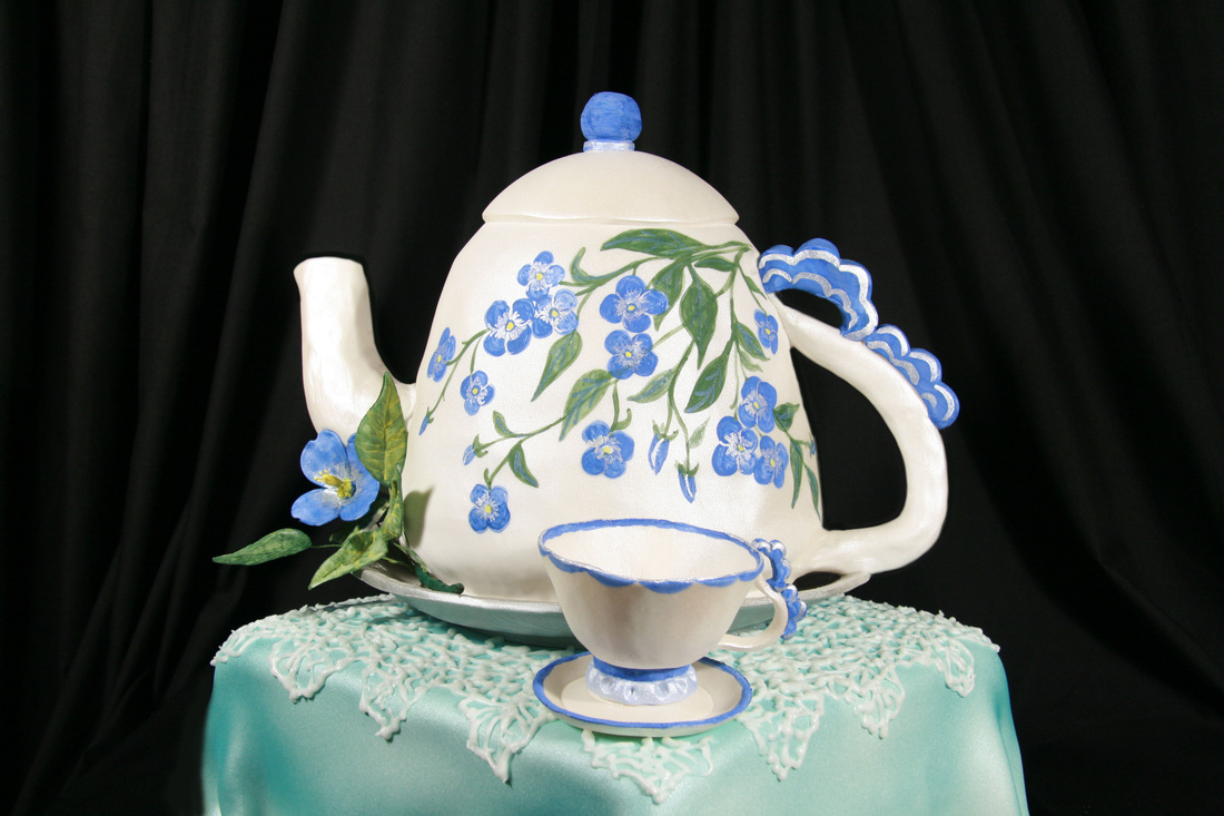 A white teapot and teacup cake decorated with blue forget-me-not flowers and green leaves sits atop a square turquoise cake base adorned with white lace piping.