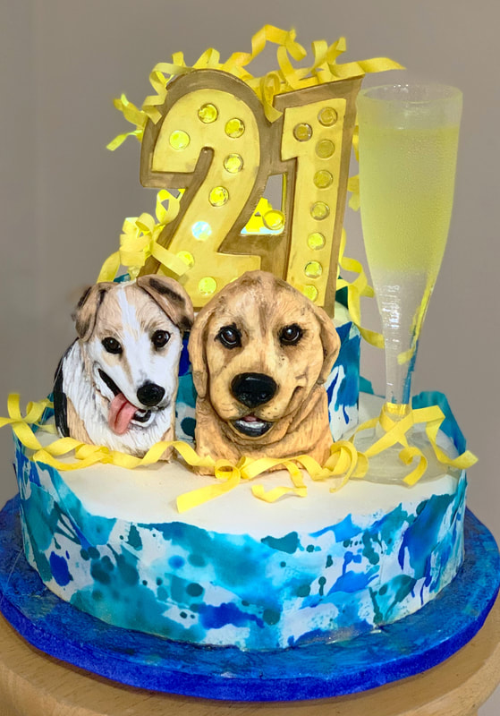 A round birthday cake with blue and white watercolor-style frosting is decorated with two sculpted dog heads, a large gold number "21" topper, yellow ribbon accents, and a fondant champagne flute.