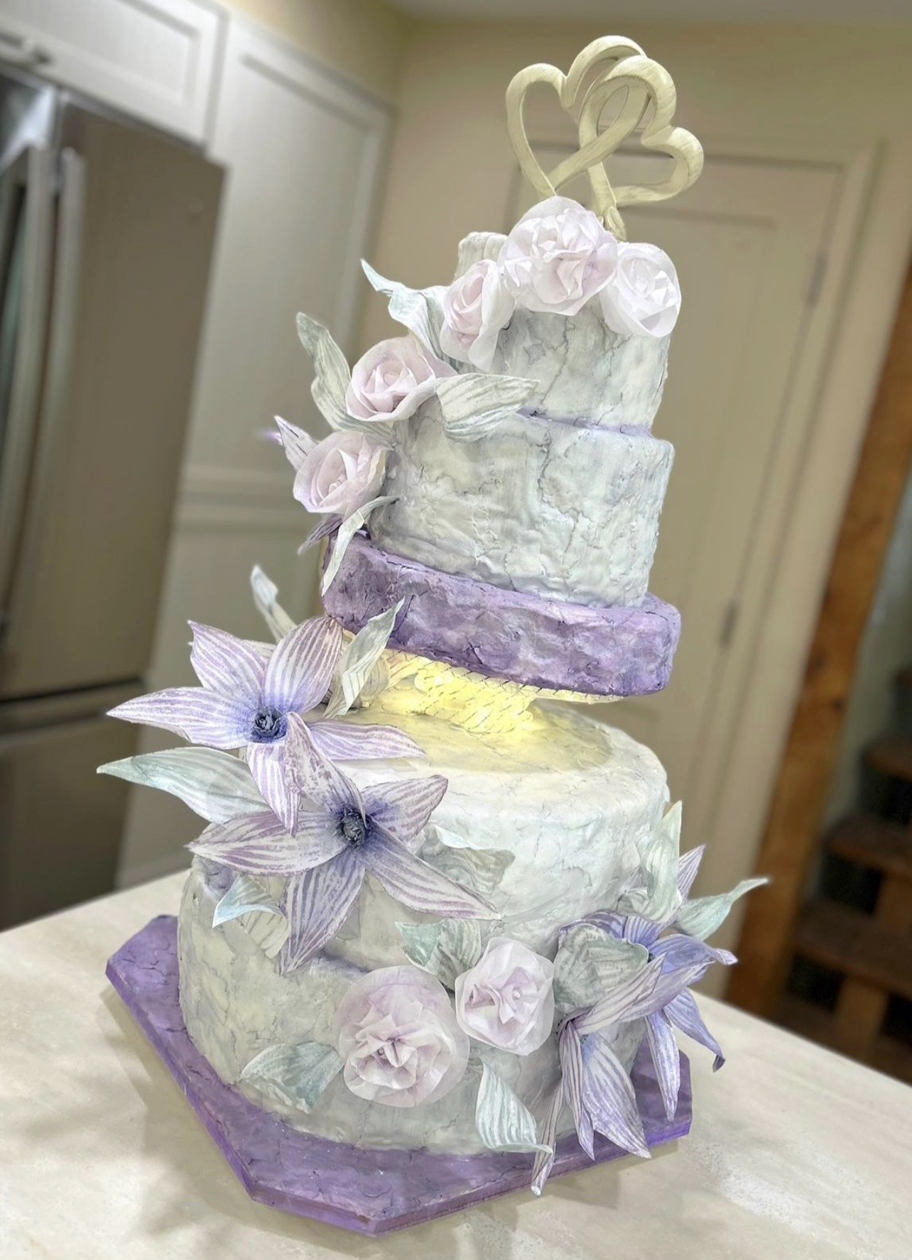A four-tiered wedding cake features a marbled fondant finish in soft pastel shades of lavender and white, each tier adorned with sugar flowers in varying shades of purple and white including roses and other blossoms interspersed with green leaves, crowned with a decorative topper of intertwined hearts and set on a square purple base.