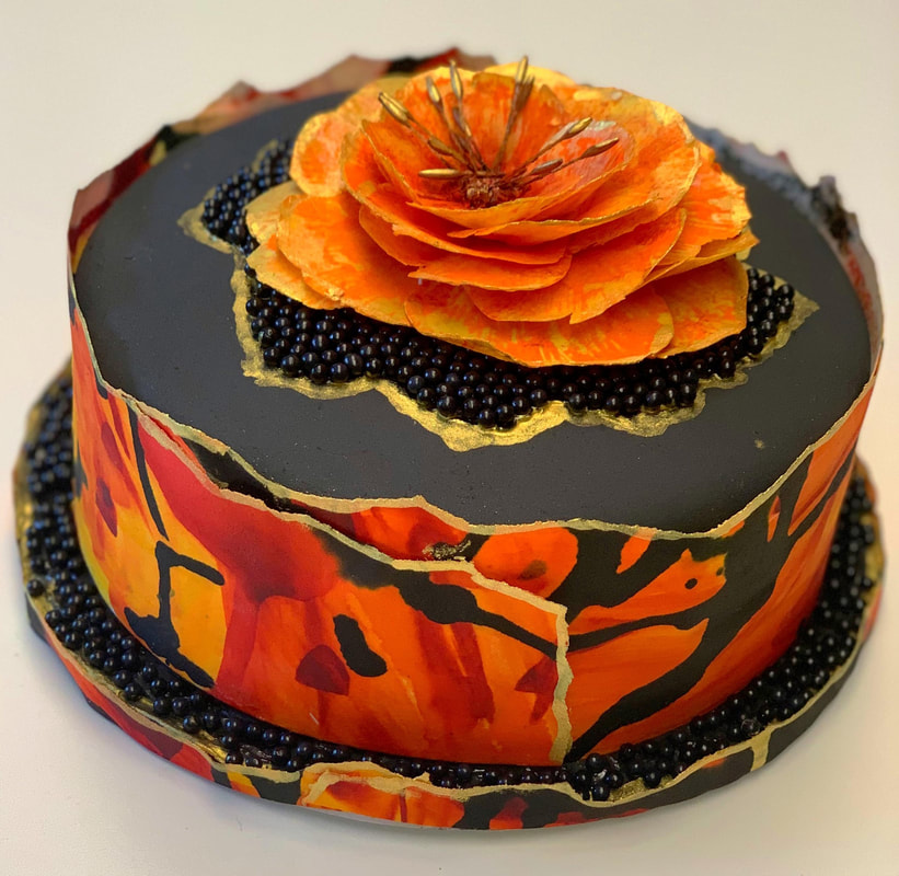 A round cake features a glossy black top and sides adorned with vibrant orange and red floral patterns, accented by gold leaf and black sugar pearls, and crowned with a large, delicate orange flower made from thin, translucent petals.
